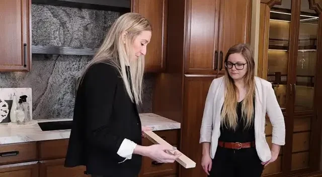 Two designers reviewing a custom woodwork sample in a showroom with rich, brown custom cabinets and detailed stone backsplash.