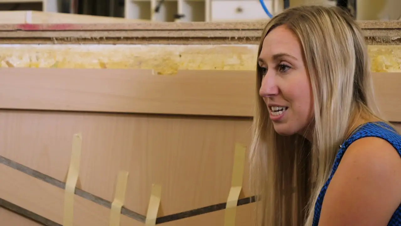A woman with long blonde hair is kneeling beside a large wooden cabinet panel in a workshop. She gestures toward the panel, which has angled wood strips held in place with yellow tape. The background shows tools and materials used in cabinetry, reinforcing the hands-on craftsmanship environment.