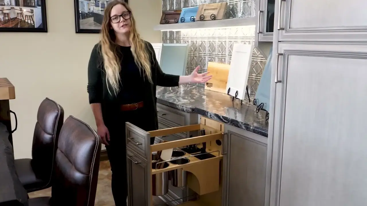 Custom kitchen cabinets with a pull-out utensil organizer displayed by a designer, showcasing detailed woodwork, soft-close drawers, and various cabinet door samples in a showroom.
