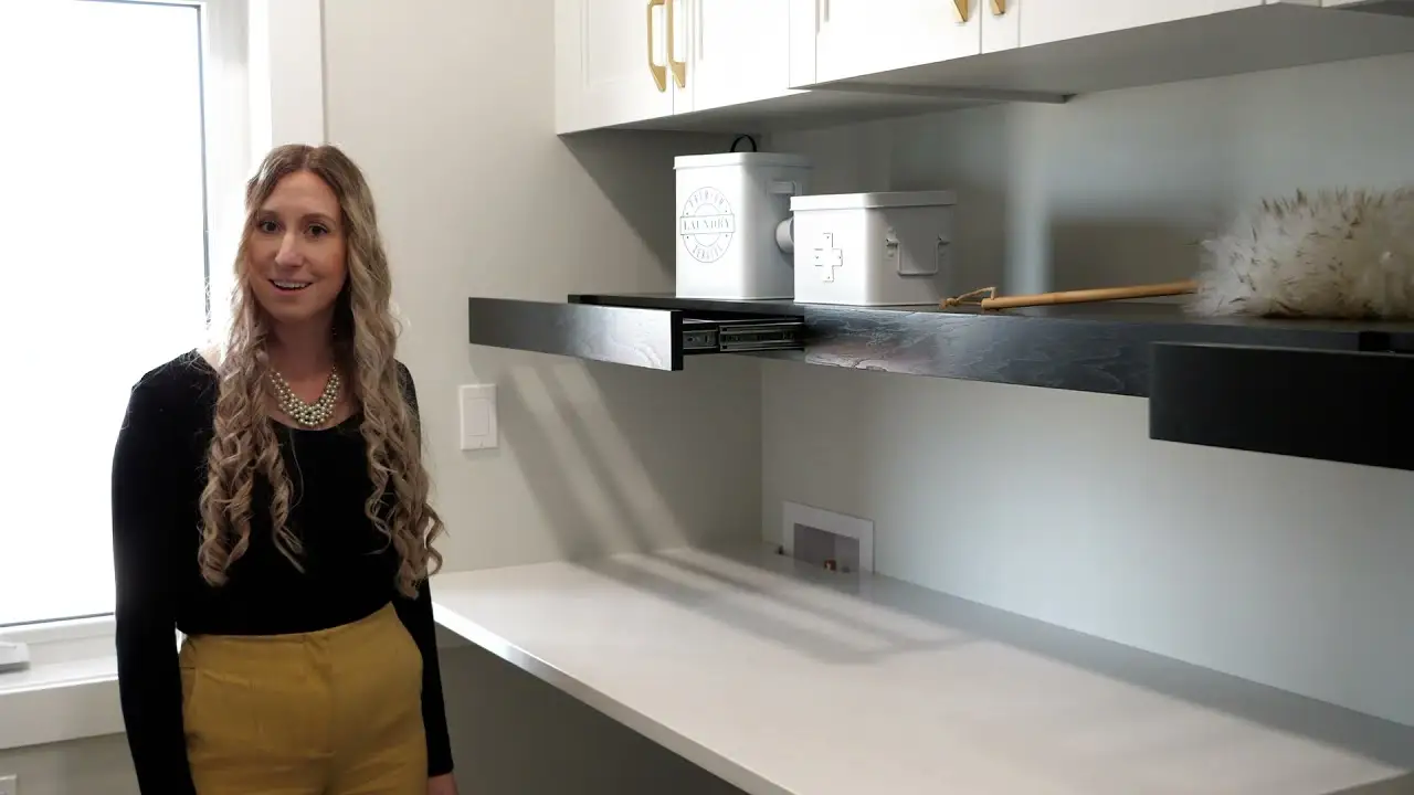 A woman with long curly hair and yellow pants stands in a modern laundry or utility room, next to a black pull-out shelf under wall cabinets.