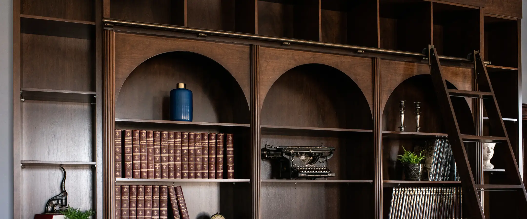 Custom dark wood built-in cabinetry with arched shelving, detailed millwork, and integrated ladder system showcasing Hiebert Cabinets’ premium craftsmanship.
