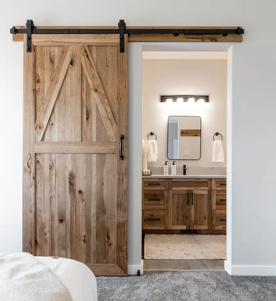 Custom rustic barn door with black hardware opening to a bathroom featuring matching wood cabinetry and detailed millwork crafted by Hiebert Cabinets.
