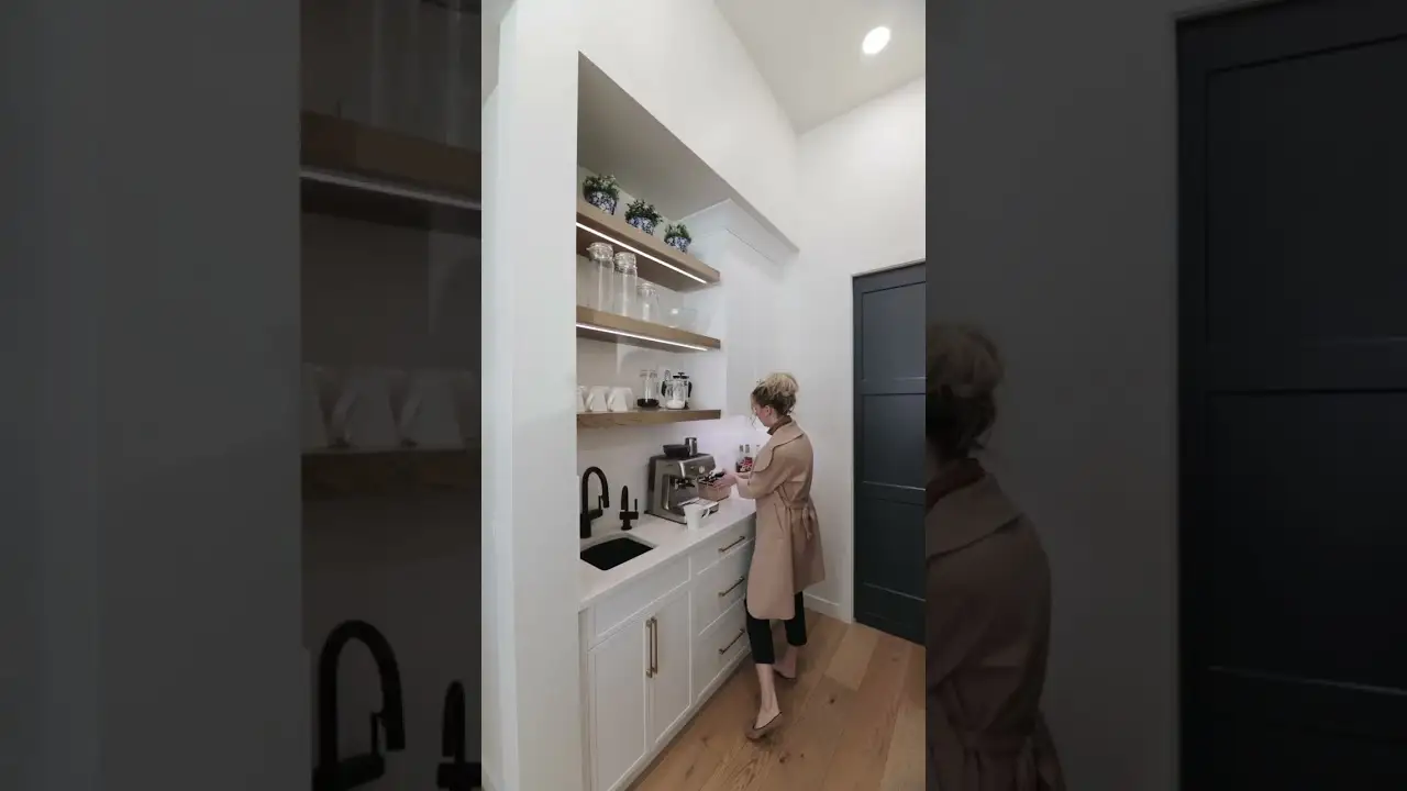 Woman standing in a compact coffee nook featuring custom white cabinets, natural wood floating shelves, and detailed millwork, showcasing high-end custom woodwork and built-in cabinetry.