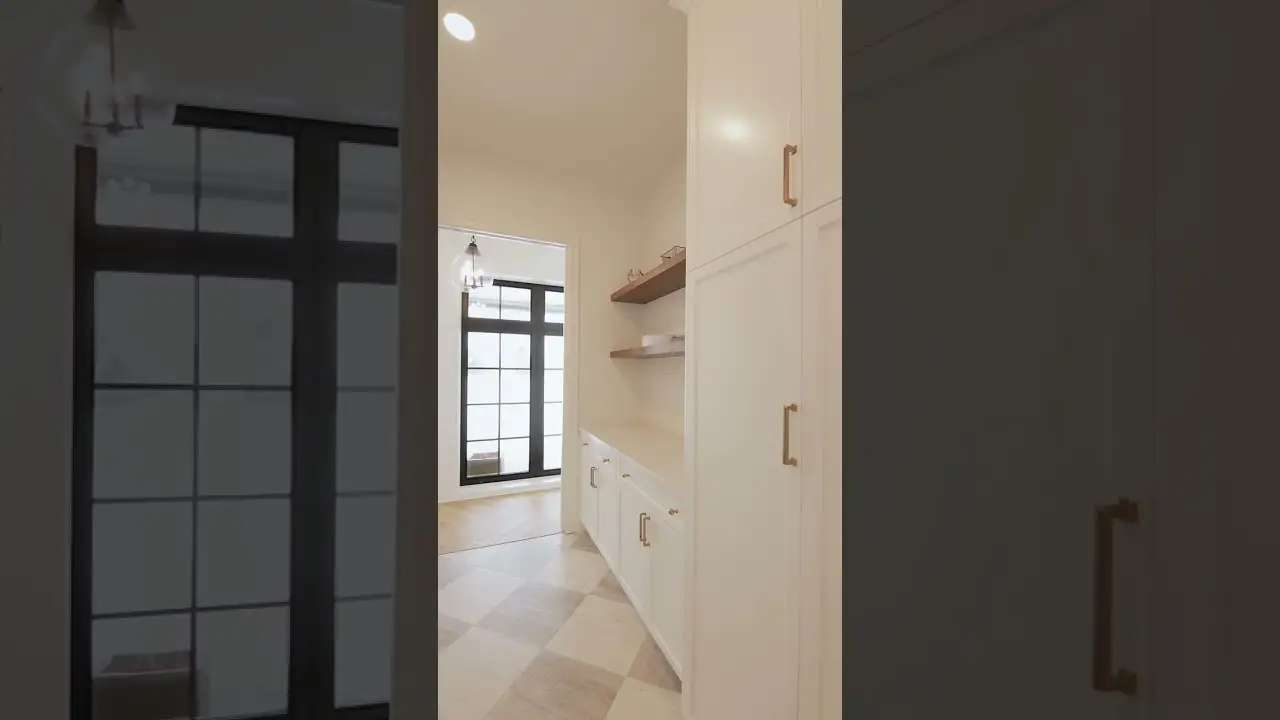 Bright hallway pantry featuring custom white cabinets with gold hardware, built-in shelving, and detailed woodwork leading into a sunlit room with large black-framed windows.