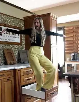 A woman standing confidently inside an open kitchen drawer in a showroom, demonstrating the strength and durability of the cabinetry. She has her arms outstretched and is smiling, showcasing the drawer’s sturdiness in a playful way.