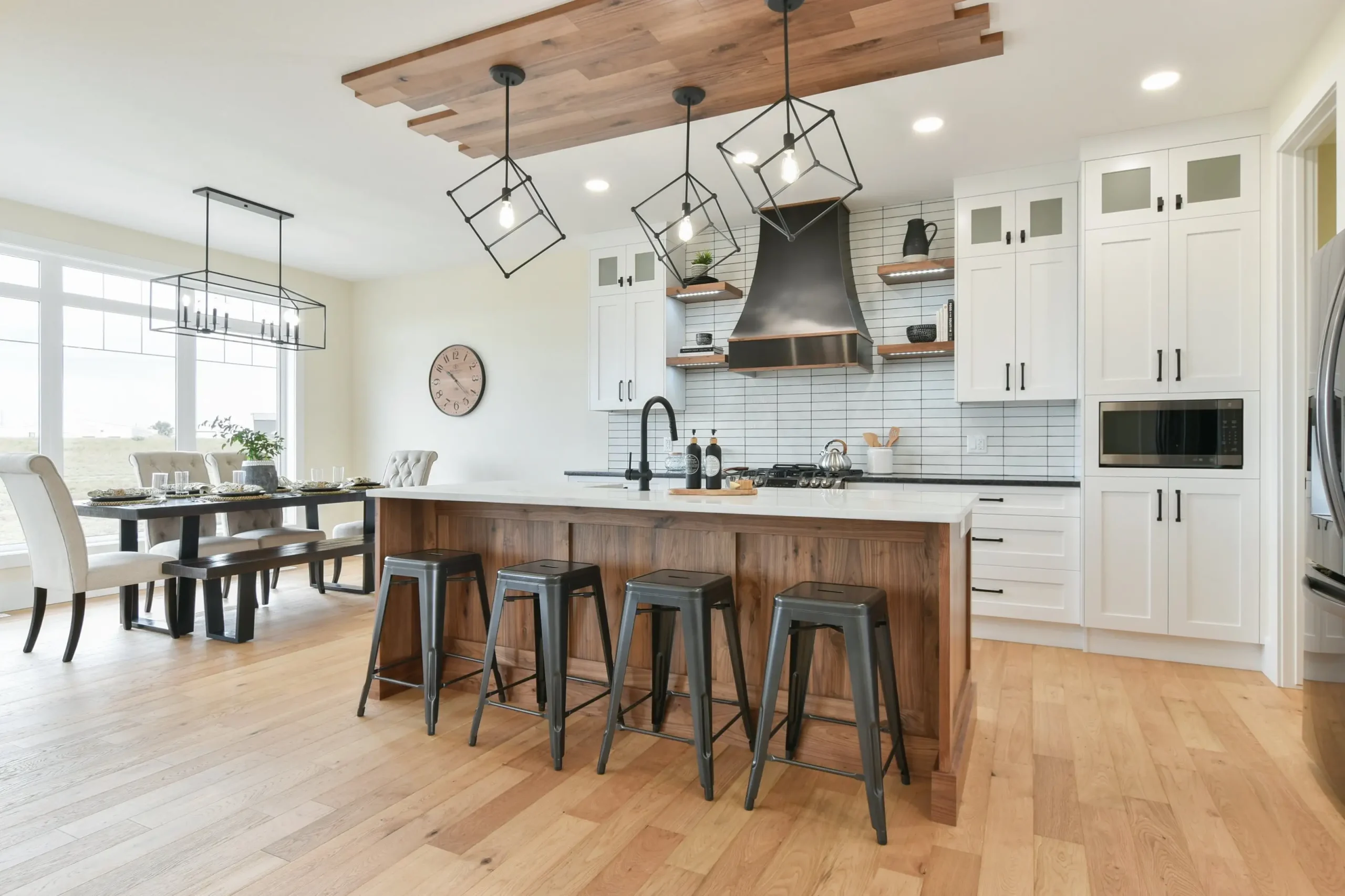 Bright modern kitchen featuring custom wood cabinets on the island, minimalist white upper cabinets, handcrafted wood hood fan detail, floating wood shelves, and a spacious dining area with natural light.