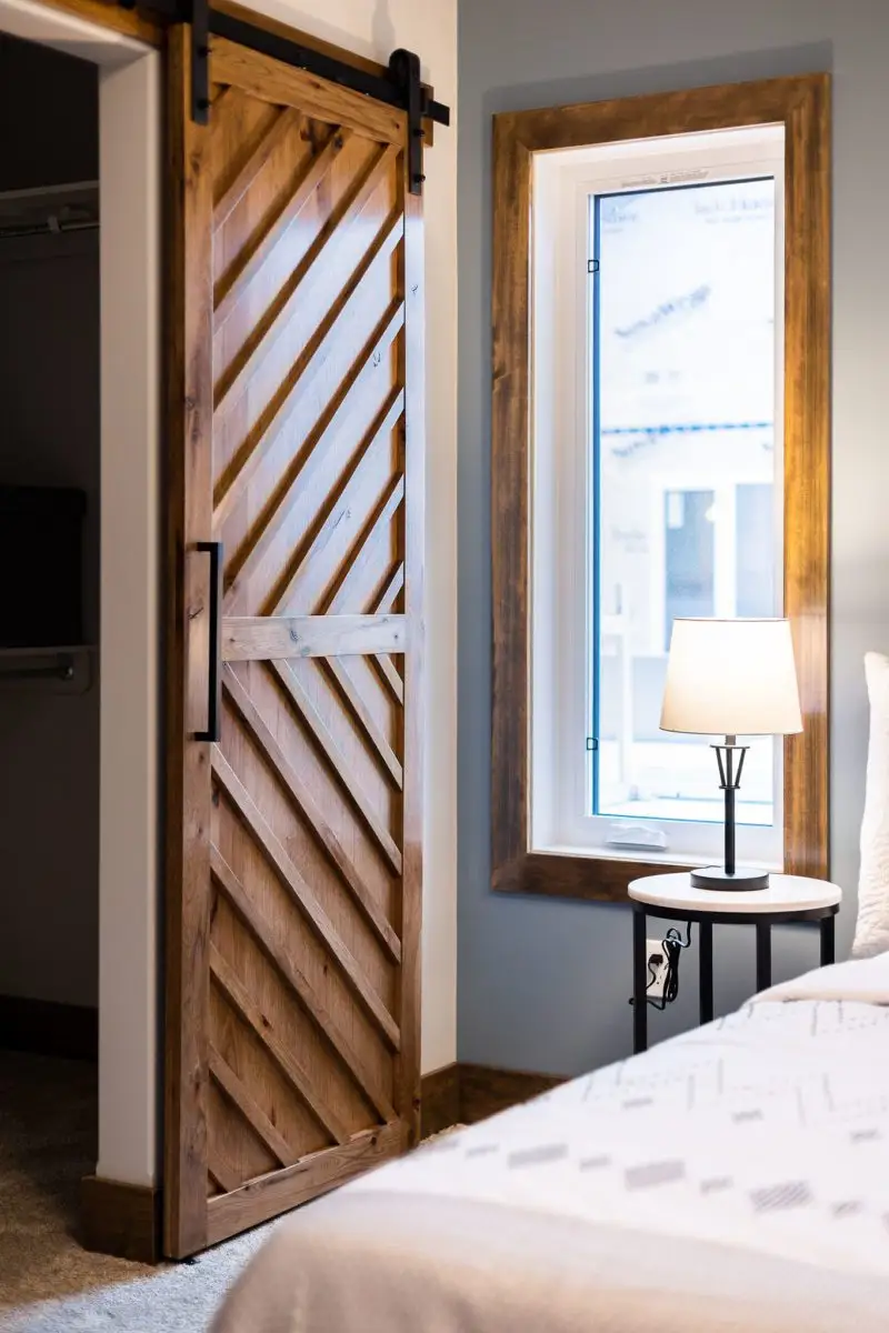 A rustic sliding barn-style wooden door with diagonal plank detailing and black metal hardware, partially open to a closet, beside a tall wood-framed window and bedside table lamp.